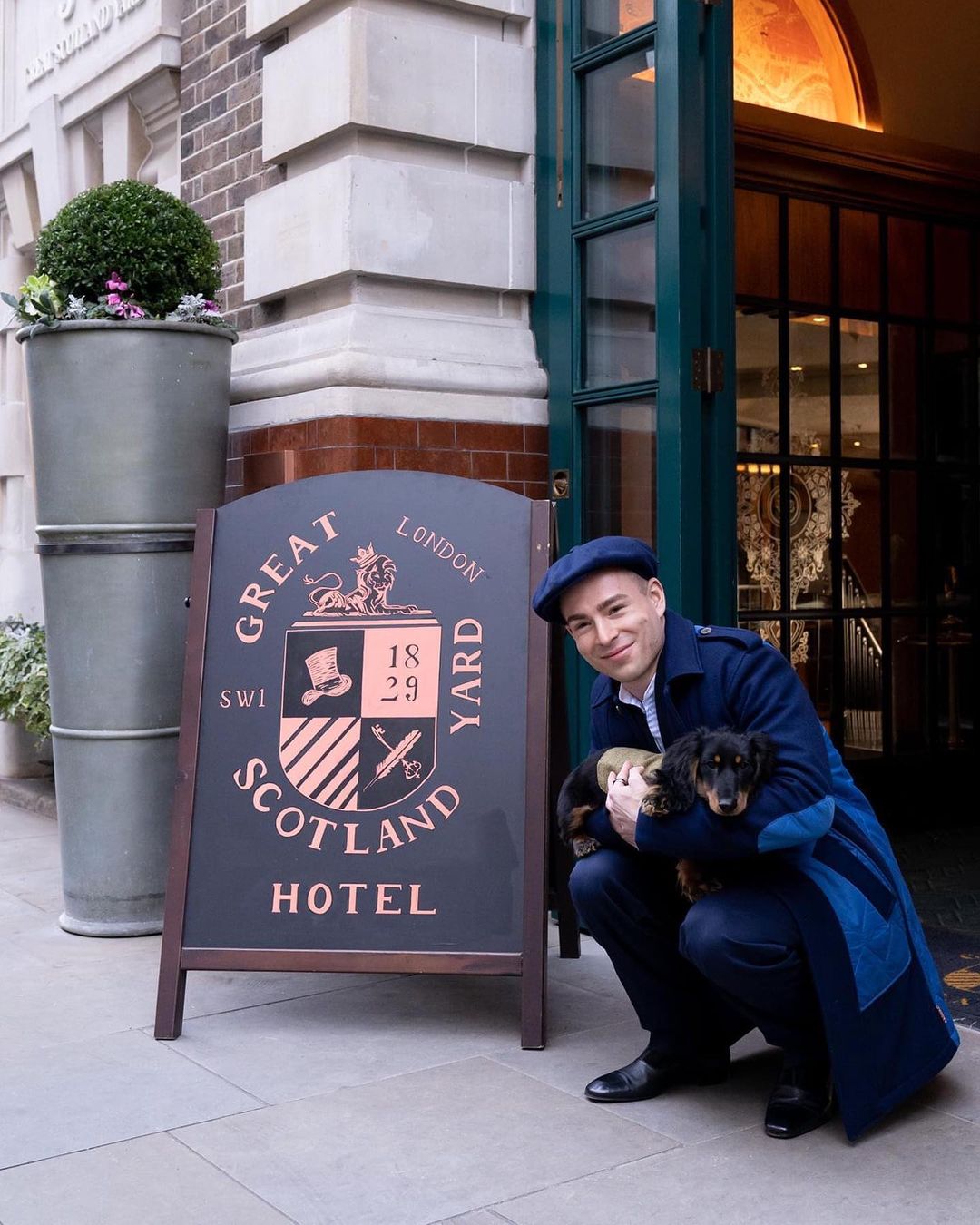 A gentleman in a blue suit is crouching next to the Great Scotland Yard Entrance, a young Dachshund in his arms. He's smiling.